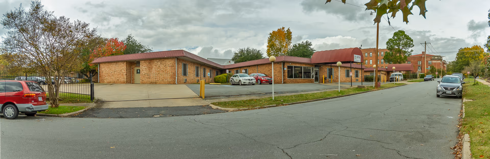 Exterior view of The Blossoms at Cumberland Rehab & Nursing Center showing a single-story brick building with a red awning over the entrance. Several cars are parked in front and along the street. Trees with autumn foliage are visible around the property under a cloudy sky.