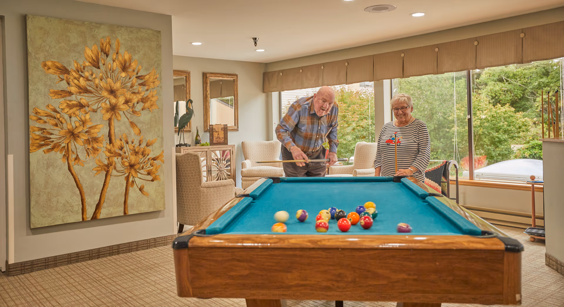 An elderly man and woman playing pool in a well-lit room with large windows showing greenery outside. The room has comfortable armchairs, a large floral painting on the wall, and a carpeted floor.