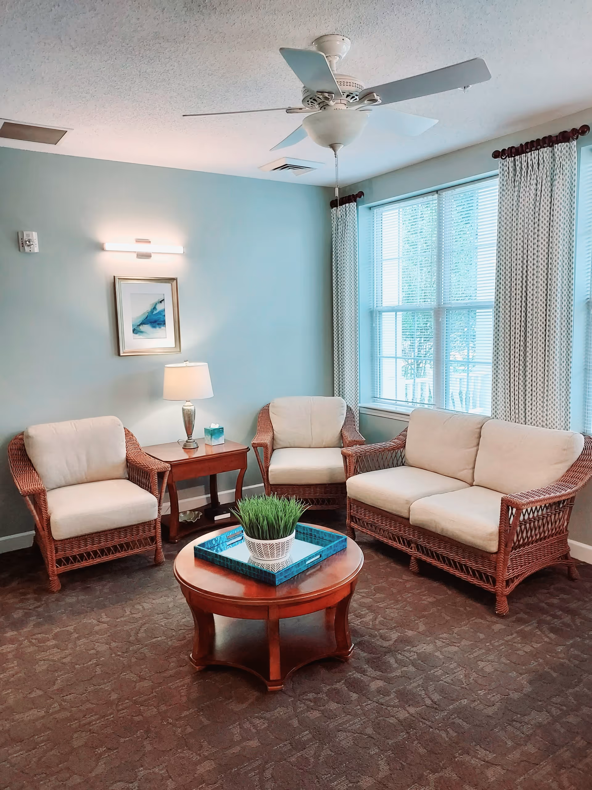 A cozy living room area with a wicker loveseat and two matching armchairs with cream cushions. A wooden coffee table with a blue tray and a potted plant sits in the center. There is a wooden side table with a lamp and a box of tissues between the chairs. The walls are painted light blue, and a framed abstract painting hangs above the side table. A large window with patterned curtains allows natural light to fill the room. A ceiling fan with light is mounted on the white ceiling.
