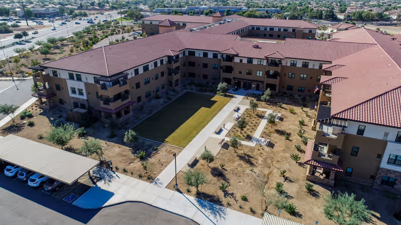 Aerial view of Clarendale Of Chandler senior living facility showing a large U-shaped building with red tile roofs surrounding a landscaped courtyard with pathways, benches, small trees, and shrubs. There is a covered parking area with several cars visible in the foreground and a suburban neighborhood in the background.