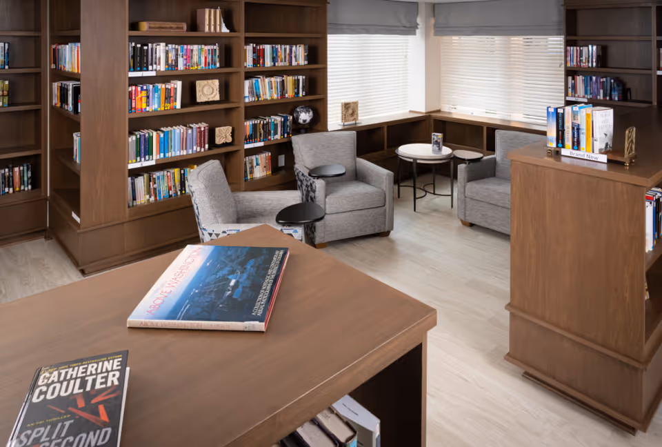 A cozy library room with wooden bookshelves filled with books, two gray armchairs with small attached tables, and round side tables near windows with blinds. A wooden table in the foreground has two books placed on it.