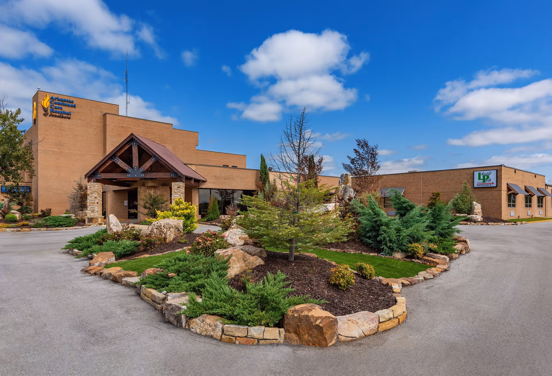 Exterior view of a large brick building with a covered entrance supported by stone pillars. There is a landscaped area with various shrubs, small trees, and rocks in front of the building. The sky is blue with scattered clouds. Signs on the building read 'Arkansas Continued Care Hospital of Jonesboro' and 'Liberty Park Assisted Living Entrance'.