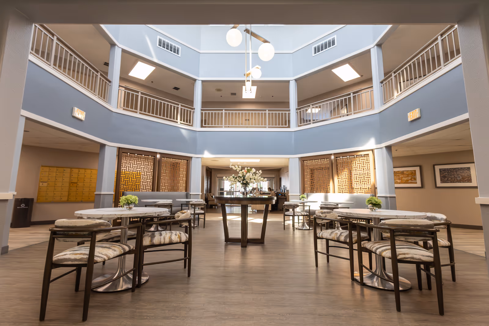 Spacious and well-lit common area in a senior living facility with round tables and cushioned chairs arranged neatly. The room features a high ceiling with modern hanging light fixtures, a second-floor balcony railing, decorative wall panels, and framed artwork on the walls. A vase with flowers is placed on a central table.