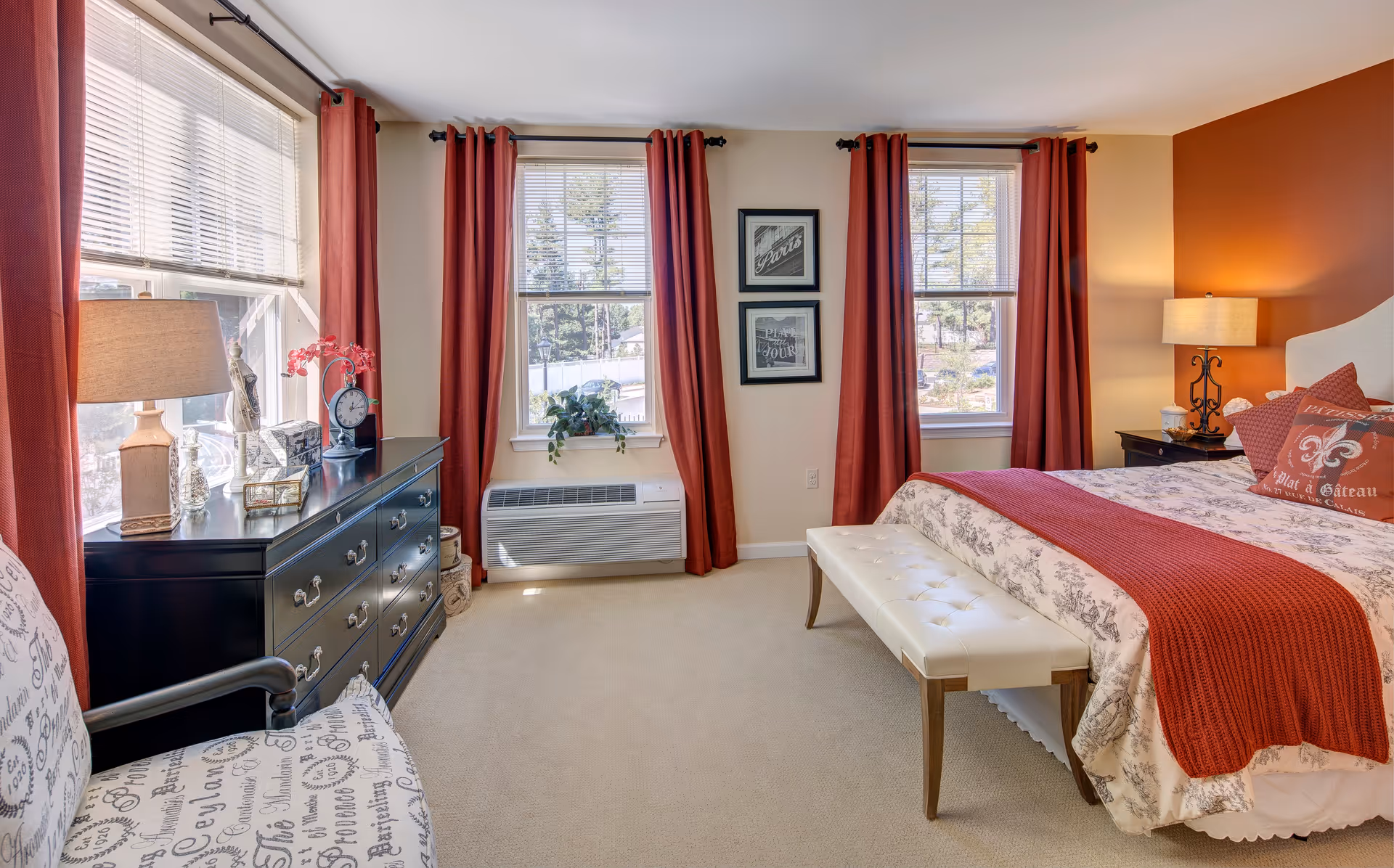 Sunlit bedroom with three windows framed by red curtains, a bed with a red throw and bench, and a black dresser with lamp.