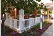 Covered front entrance of a brick senior living building with a white porch railing, columns, landscaping and a sidewalk leading to a sign.