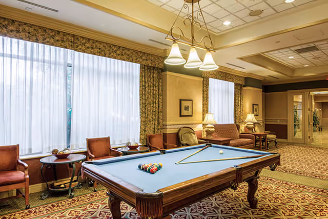 A well-lit common area featuring a pool table with balls and cues arranged on it, surrounded by chairs and a sofa. The room has large windows with sheer curtains, patterned carpet, and warm lighting from ceiling fixtures and table lamps.