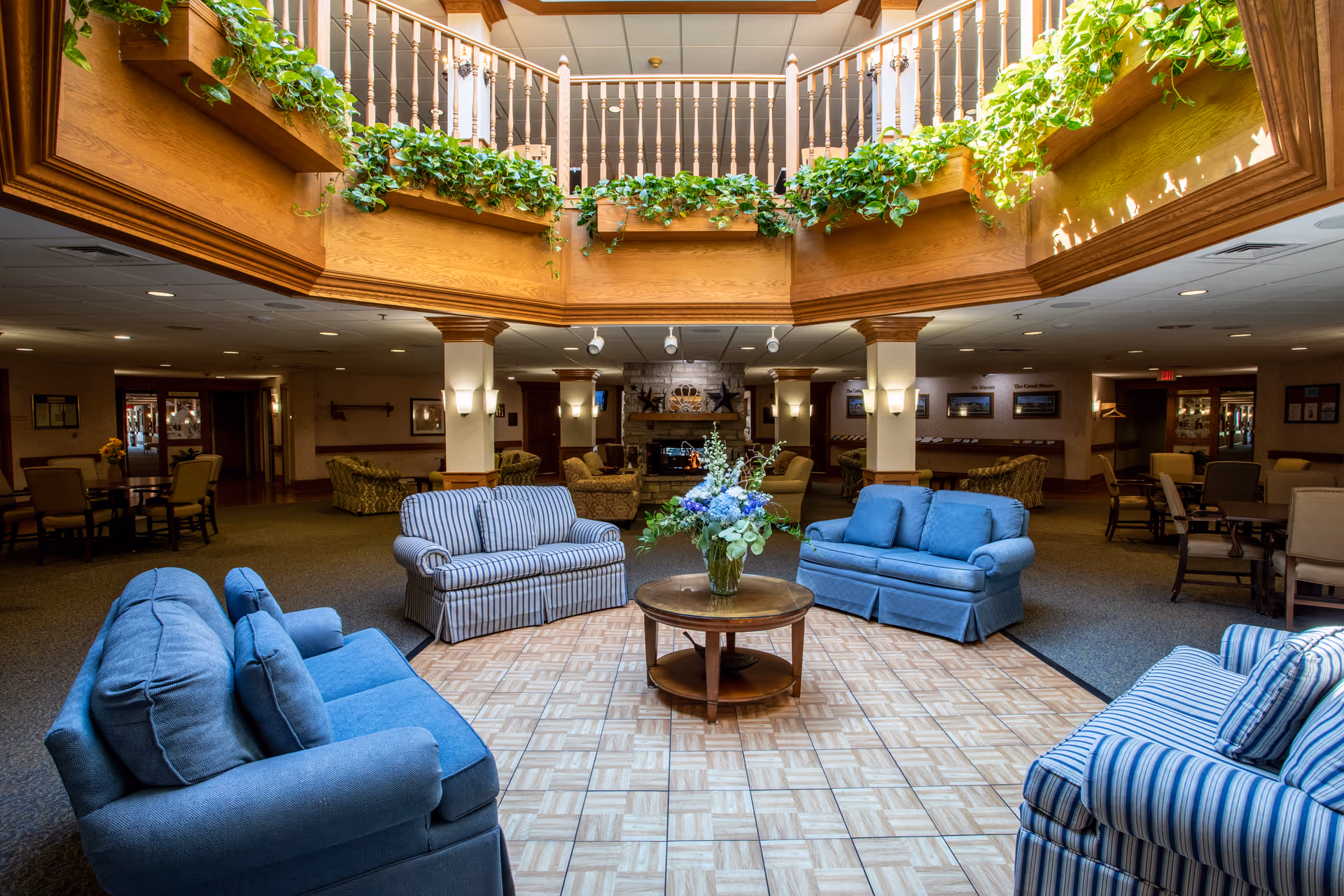 A bright senior living facility lounge with blue sofas arranged around a central table under a skylight with hanging plants.