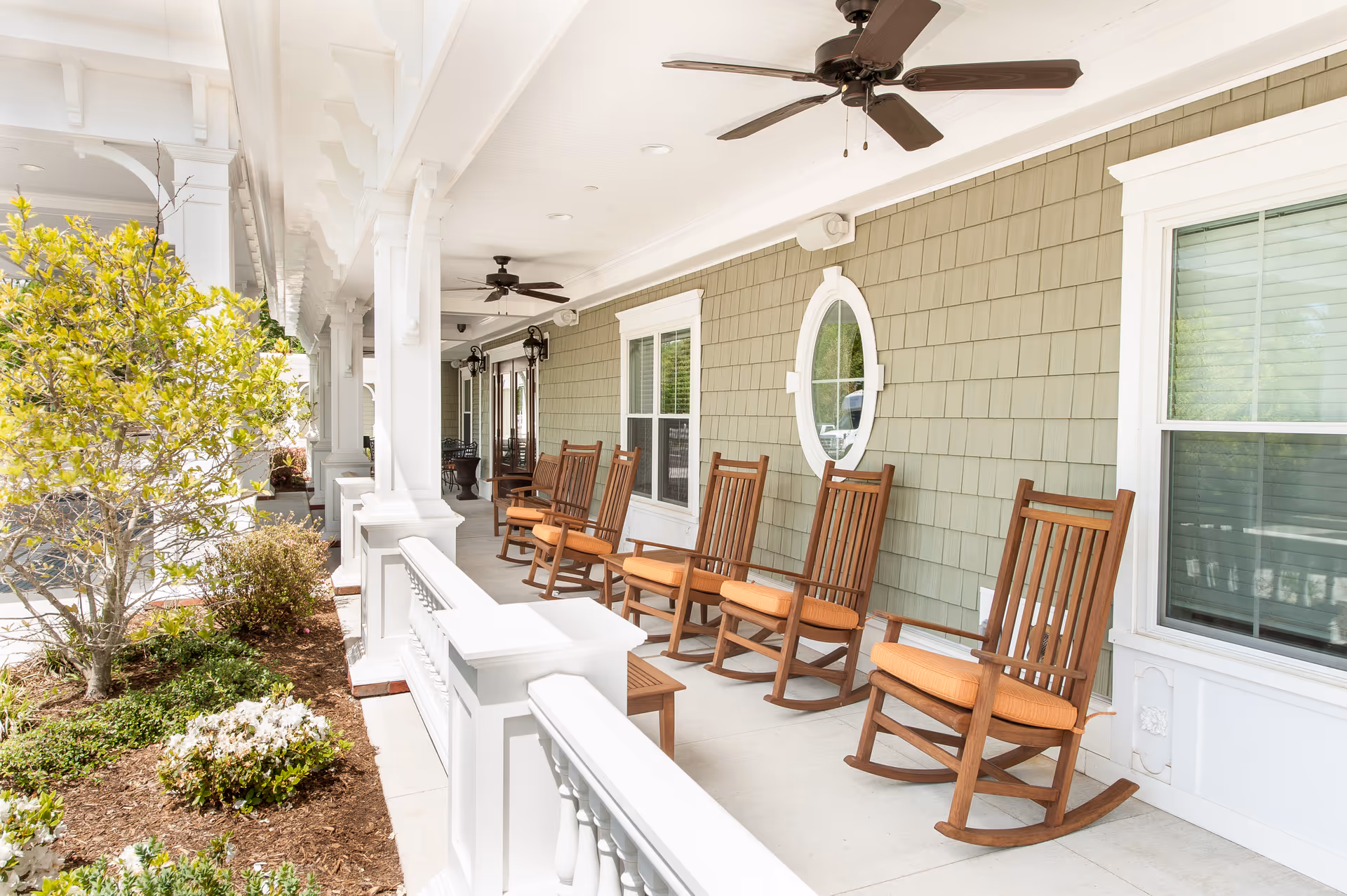 A covered porch area with several wooden rocking chairs with orange cushions lined up against a green shingled wall. The porch has white railings and columns, ceiling fans, and windows with white trim. There are plants and shrubs visible next to the porch.