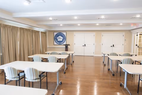 A bright room with several white tables and striped chairs arranged in rows on a wooden floor. Beige walls and large beige curtains cover the windows on the left side. There are double white doors at the far end of the room and a blue and white emblem on the wall.