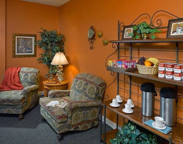 A cozy sitting area with two patterned armchairs, one with an open book on it and the other with a red throw blanket. A small round wooden table with a lamp and a decorative plant is placed between the chairs. On the right side, a metal shelving unit holds coffee dispensers, cups, a basket of fruit, canned goods, and some framed pictures. The walls are painted a warm orange color.