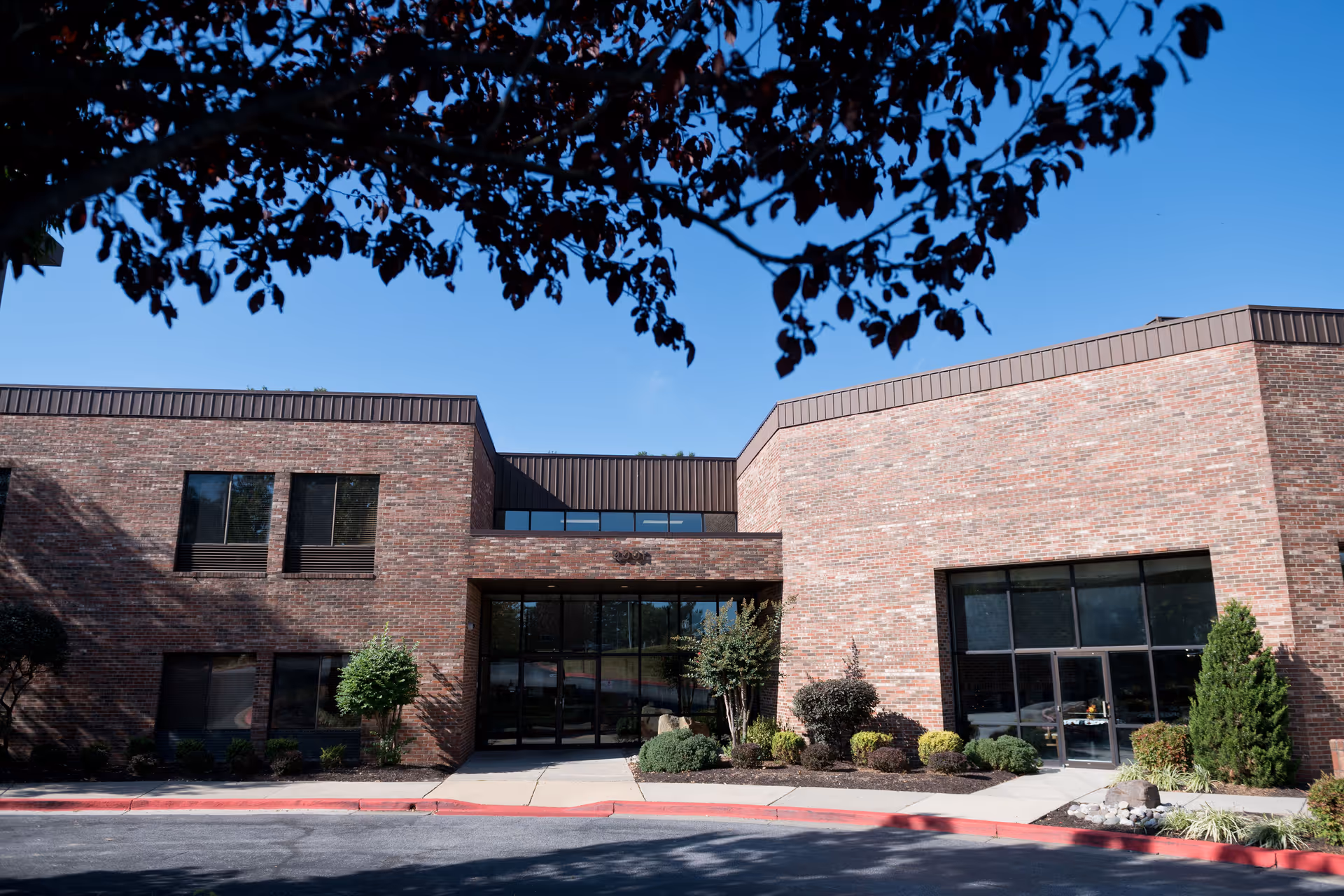 Front exterior view of a brick building with large windows and a glass entrance door, surrounded by small shrubs and trees, under a clear blue sky with tree branches partially framing the top of the image.