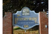 A blue and gold sign mounted on two brick pillars that reads 'The Bickford Home N.Y.S. Certified Home for Adults 56' with a decorative emblem featuring a horse and rider at the top, set outdoors with trees and grass in the background.