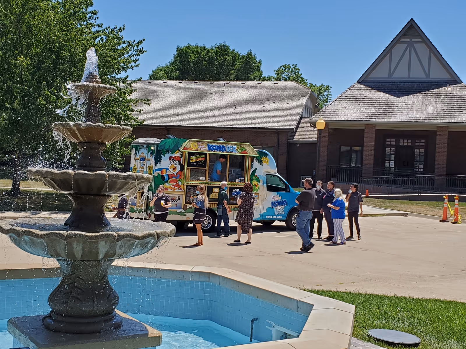 A three-tiered fountain in a courtyard with a colorful Kona Ice truck and several people gathered outside a brick building.