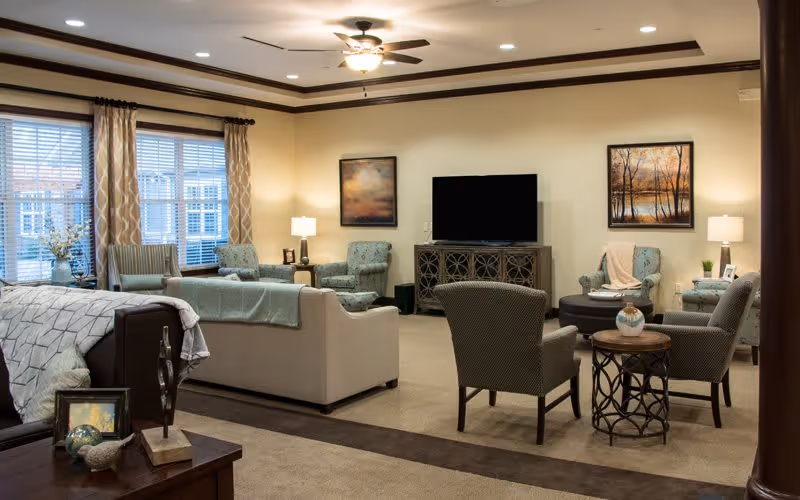 A spacious and well-lit living room with multiple armchairs and sofas arranged around a central area. There is a large flat-screen TV on a decorative console against the wall, flanked by two framed landscape paintings. The room features beige walls, carpeted floors, a ceiling fan with lights, and large windows with patterned curtains allowing natural light to enter. Several side tables hold lamps and decorative items.