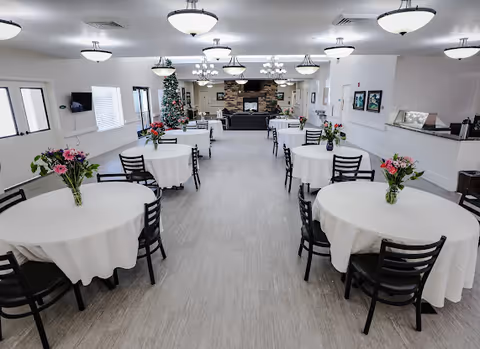 Bright communal dining room with round tables covered in white tablecloths and floral centerpieces, chandeliers overhead, and a fireplace with a Christmas tree at the far end.