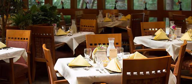 Dining room with multiple tables set with white tablecloths, folded yellow napkins, glassware and wooden chairs.