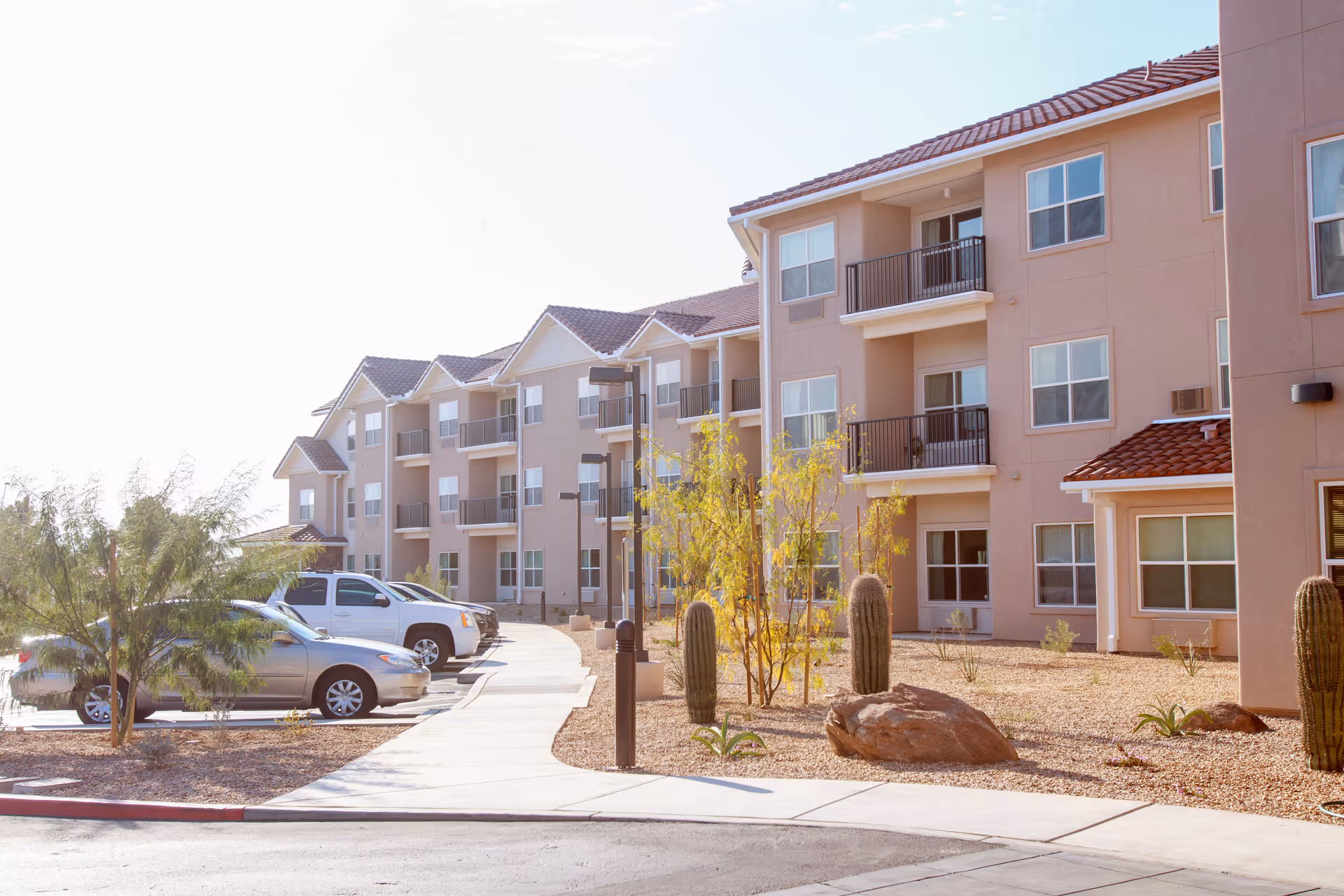 Exterior view of a three-story senior living facility building with beige walls and red-tiled roofs. There are balconies on the second and third floors. In front of the building, there is a landscaped area with desert plants including cacti and small trees, as well as a sidewalk and a parking lot with several parked cars.