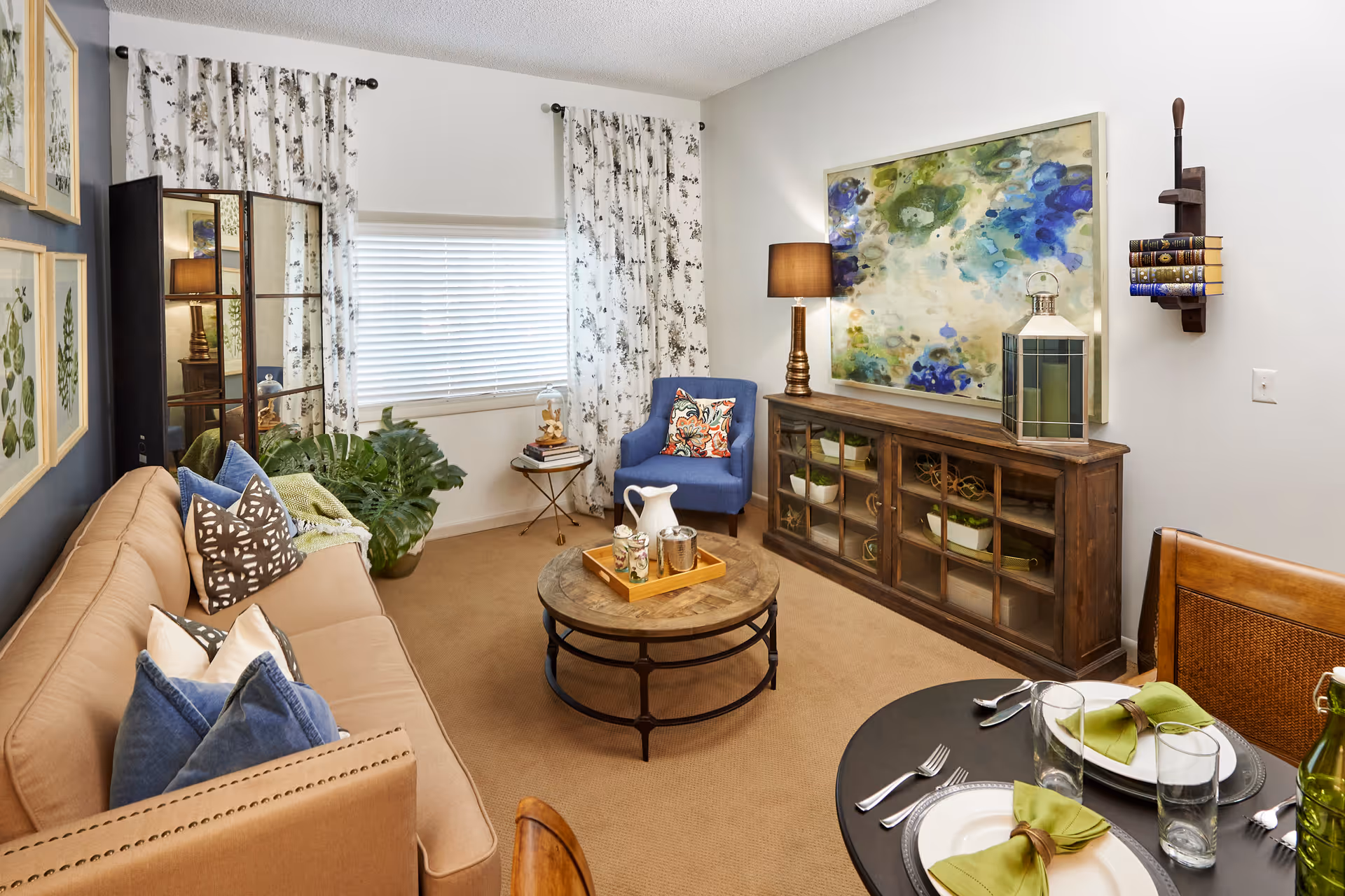 A cozy living room with a beige sofa adorned with multiple pillows, a round wooden coffee table with a tray holding a pitcher and cups, a blue armchair with a colorful pillow, a wooden cabinet with glass doors, a large abstract painting on the wall, a table lamp, and a small round dining table set with plates, glasses, and green napkins. The room has floral curtains and a window with blinds.