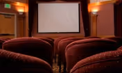 Interior view of a small theater or screening room with rows of red upholstered seats facing a large white projection screen. The room has warm lighting and wood-paneled walls.