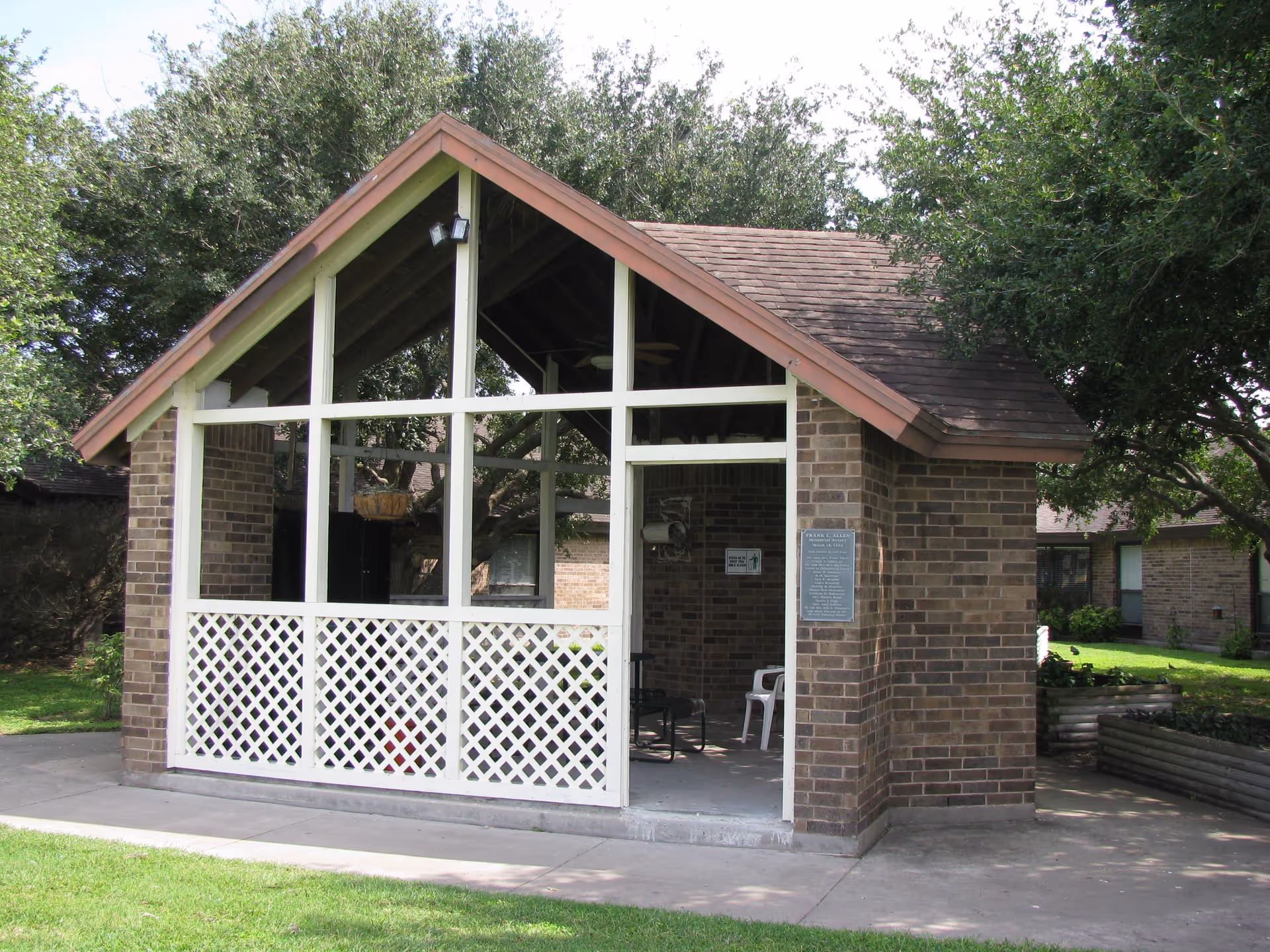 A small brick pavilion with a peaked roof and white lattice panels on the lower half of the front side. The pavilion is situated outdoors on a concrete path surrounded by green grass and trees. Inside, there are a few chairs and a hanging basket. The setting appears to be part of a senior living facility.