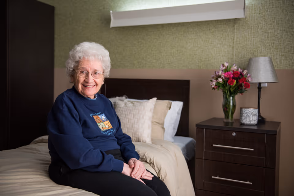 An elderly woman with white hair and glasses is sitting on a bed in a bedroom. She is smiling and wearing a navy blue sweatshirt. The bed has beige and white pillows and bedding. Next to the bed is a dark wooden nightstand with a vase of colorful flowers, a lamp, and a decorative candle. The wall behind the bed has a green textured wallpaper with a beige lower section.