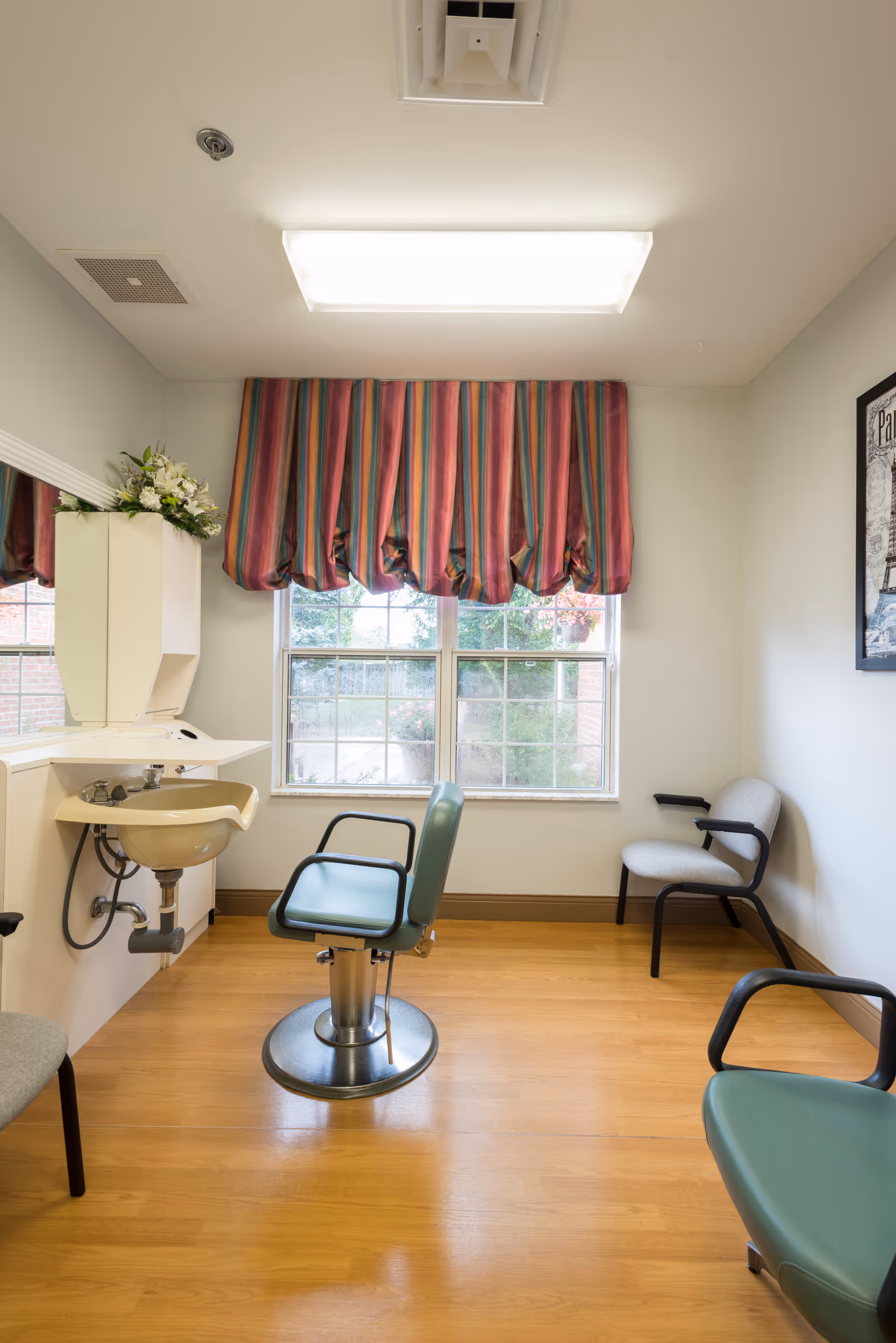 A small room with a salon-style chair in front of a sink and mirror, two additional chairs along the walls, a window with colorful striped curtains, and a framed picture on the wall.