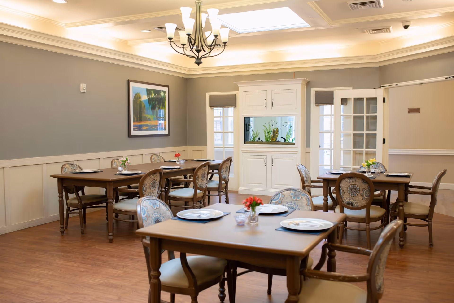 A well-lit dining room with wooden tables and upholstered chairs arranged neatly. Each table has plates and small flower vases as centerpieces. The room features a chandelier, a built-in white cabinet with an aquarium, and framed artwork on the wall. The floor is wooden, and there are windows with blinds allowing natural light.