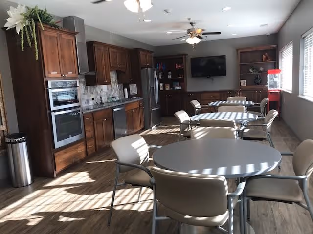 A bright room with wooden cabinetry and kitchen appliances including an oven, dishwasher, and refrigerator. Several round tables with beige chairs are arranged in the room, which has wood flooring and large windows letting in natural light. Ceiling fans and a wall-mounted TV are visible, along with a popcorn machine in the corner.