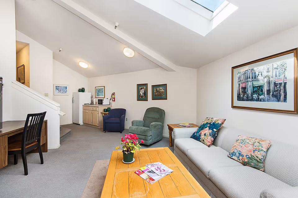 A bright living room area with a white couch adorned with floral cushions, a wooden coffee table with magazines and a potted plant, two armchairs, a small kitchenette with a refrigerator, microwave, and cabinets, and framed artwork on the walls. There is a skylight on the ceiling allowing natural light to fill the room.