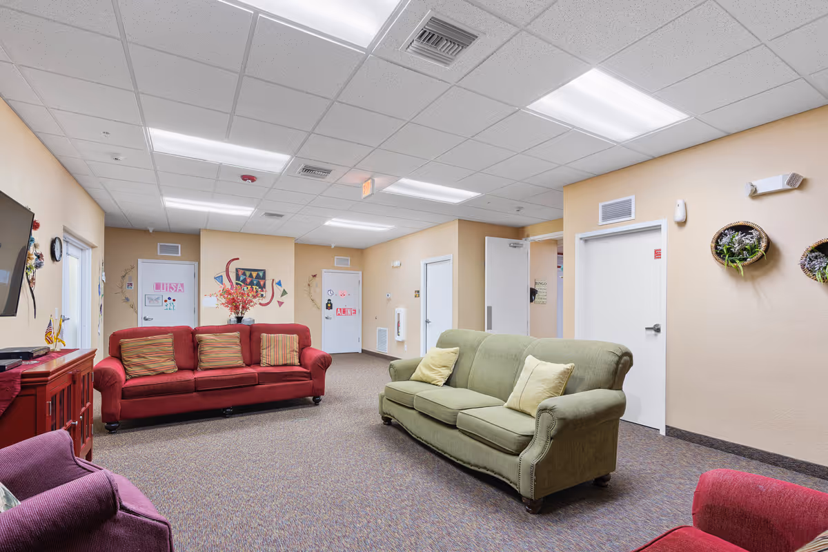A well-lit assisted living facility common area with a green sofa and a red sofa, each with cushions. The walls are beige with decorative wall hangings and doors labeled with names. There is a red cabinet with a TV mounted above it on the left side of the room.