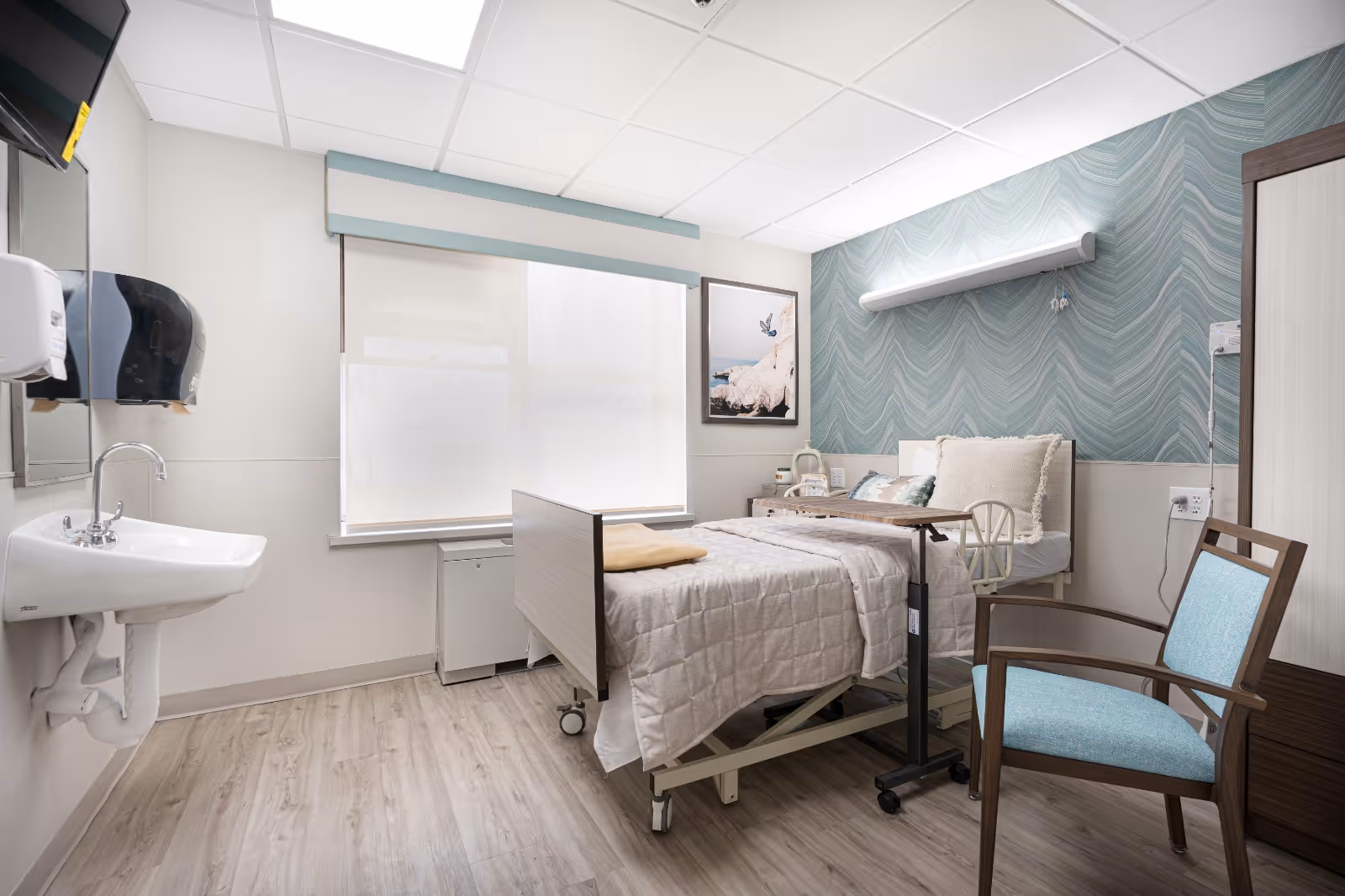 A clean and bright healthcare facility bedroom with a hospital bed covered in a beige quilt, a light blue cushioned chair, a small overbed table, a wall-mounted sink with soap dispensers, a window with a white roller shade, and a decorative blue patterned accent wall behind the bed.