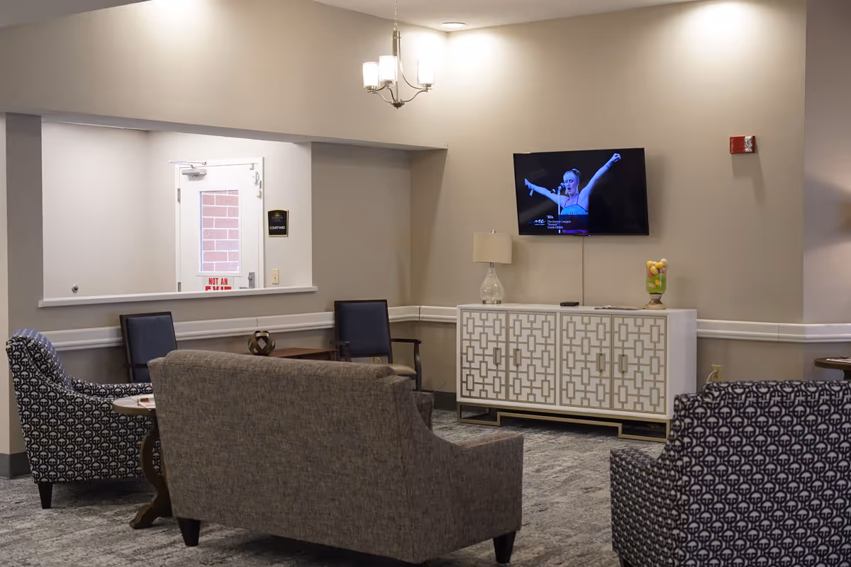 A cozy living room area with a gray sofa and two patterned armchairs arranged around a small wooden table. A flat-screen TV is mounted on the beige wall above a white cabinet with a geometric pattern. A table lamp and a decorative glass container with fruit are on the cabinet. The room has a carpeted floor and neutral-colored walls with a chandelier hanging from the ceiling.
