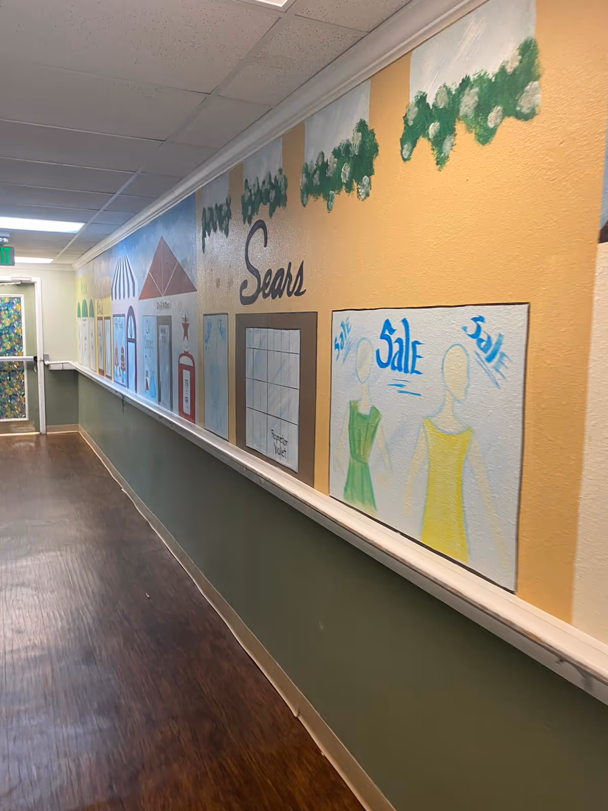 A hallway with a mural painted on the right wall depicting storefronts and sale signs, including a Sears store. The floor is wooden, the lower half of the wall is painted green, and the ceiling has white tiles. A door with a colorful pattern is visible at the end of the hallway.
