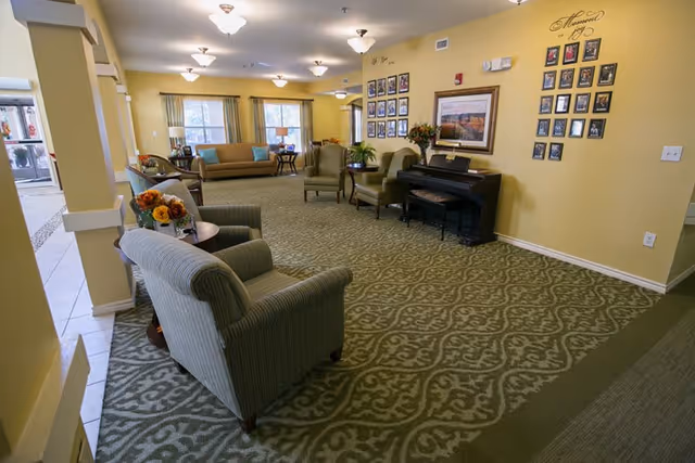 A spacious senior living facility common area with patterned carpet, yellow walls, and multiple seating options including armchairs and sofas. There is a piano against the wall with framed photos and artwork above it. The room is well-lit with ceiling lights and natural light from windows.