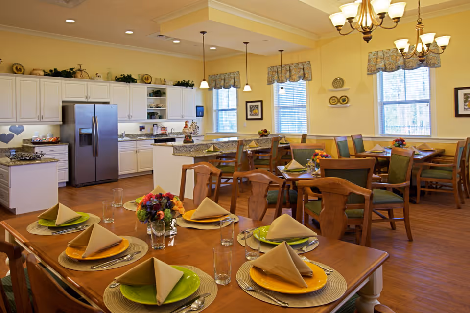 A bright and cozy dining room with wooden tables and chairs set with colorful plates, folded napkins, glasses, and floral centerpieces. The room features large windows with valances, warm yellow walls, and a kitchen area with white cabinets and stainless steel refrigerator in the background. Pendant and chandelier lighting illuminate the space.