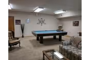 A cozy recreation room with a pool table in the center, surrounded by upholstered chairs and a sofa. The walls are decorated with a clock, a metal wall art piece, and framed pictures. The room is carpeted and has fluorescent ceiling lights.