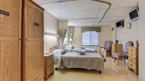 A hospital-style patient room with beds, chairs, and wooden furniture.