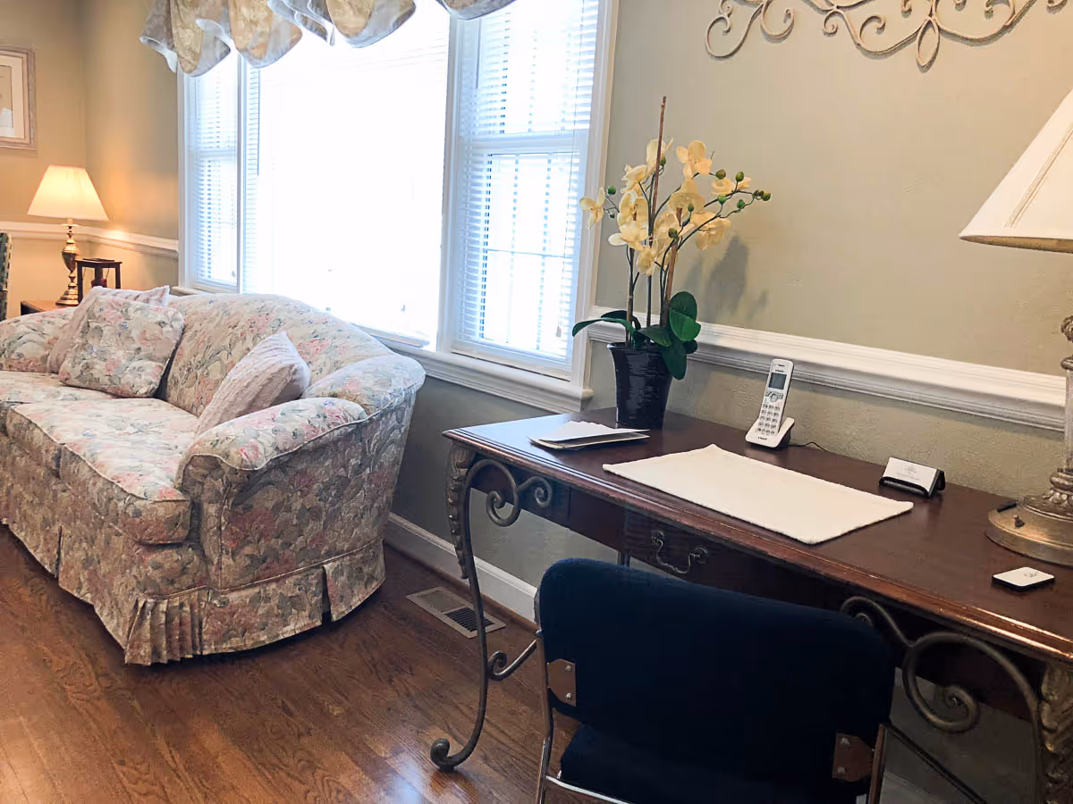 A cozy living room area with a floral-patterned sofa, a wooden desk with a white runner, a cordless phone, a potted orchid plant, and a table lamp. The room has large windows with white blinds and light-colored walls with decorative molding.