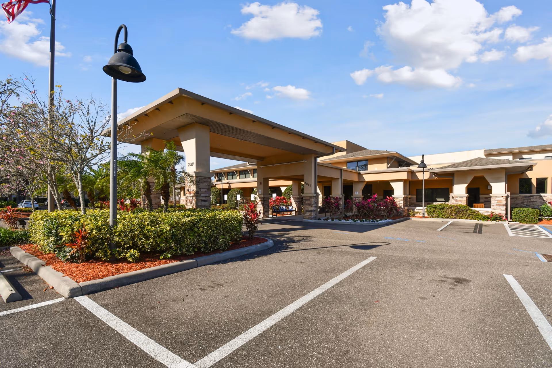 Front entrance of The Preserve at Dunedin with a covered drop-off canopy, landscaped beds and empty parking spaces under a blue sky.