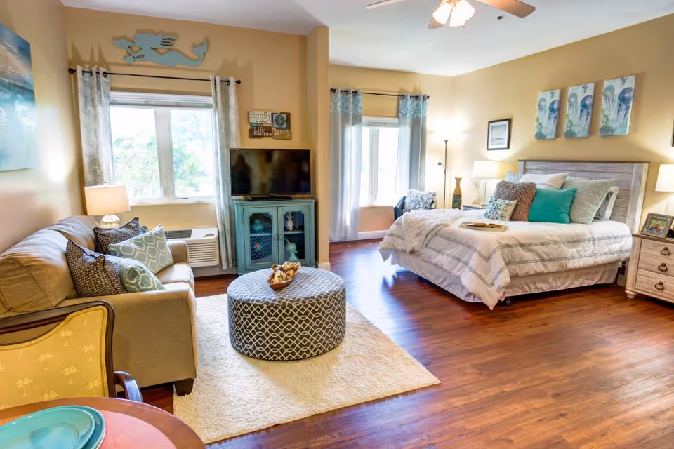 Bright studio-style senior living interior with a bed, seating area, TV console, and hardwood floors.
