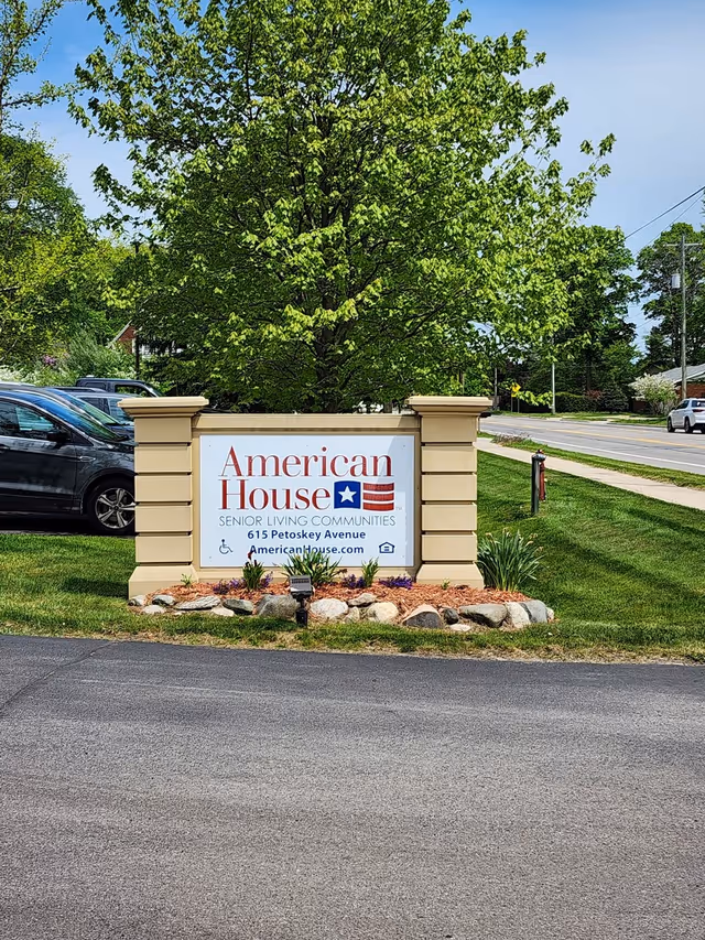 Entrance sign for American House senior living community with landscaping, parked cars, and a tree-lined street.