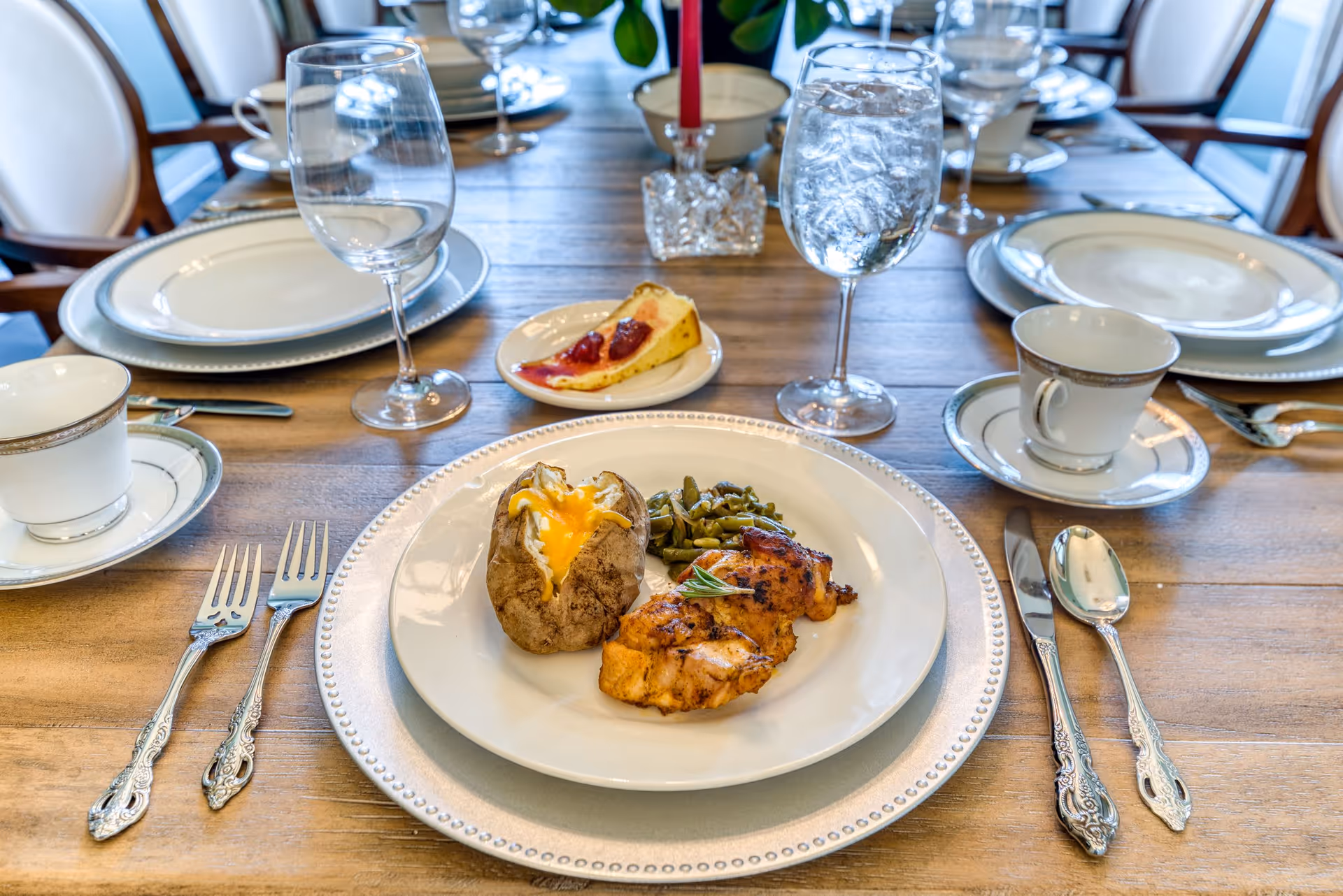 A dining table set with plates, silverware, wine glasses, and cups. The main plate in the foreground contains a baked potato with melted cheese, grilled chicken, and green beans. A slice of pie is on a small plate behind the main plate. The table is wooden and surrounded by chairs.