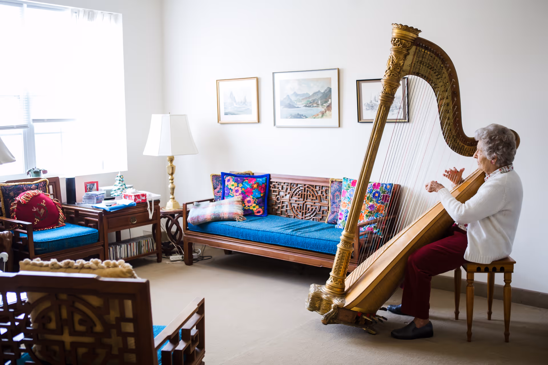An elderly woman playing a large golden harp while seated on a wooden stool in a bright living room. The room features wooden furniture with blue cushions and colorful pillows, a side table with a lamp, and framed artwork on the white walls.