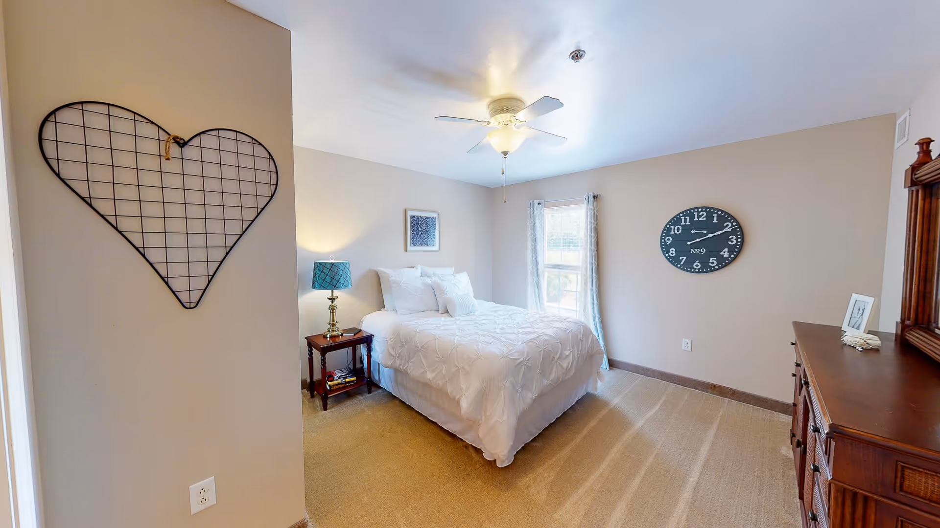 A cozy bedroom with a white bedspread on a double bed, a wooden nightstand with a blue lamp, a window with light curtains, a large wall clock, a wooden dresser with a mirror, and a decorative wire heart hanging on the wall.