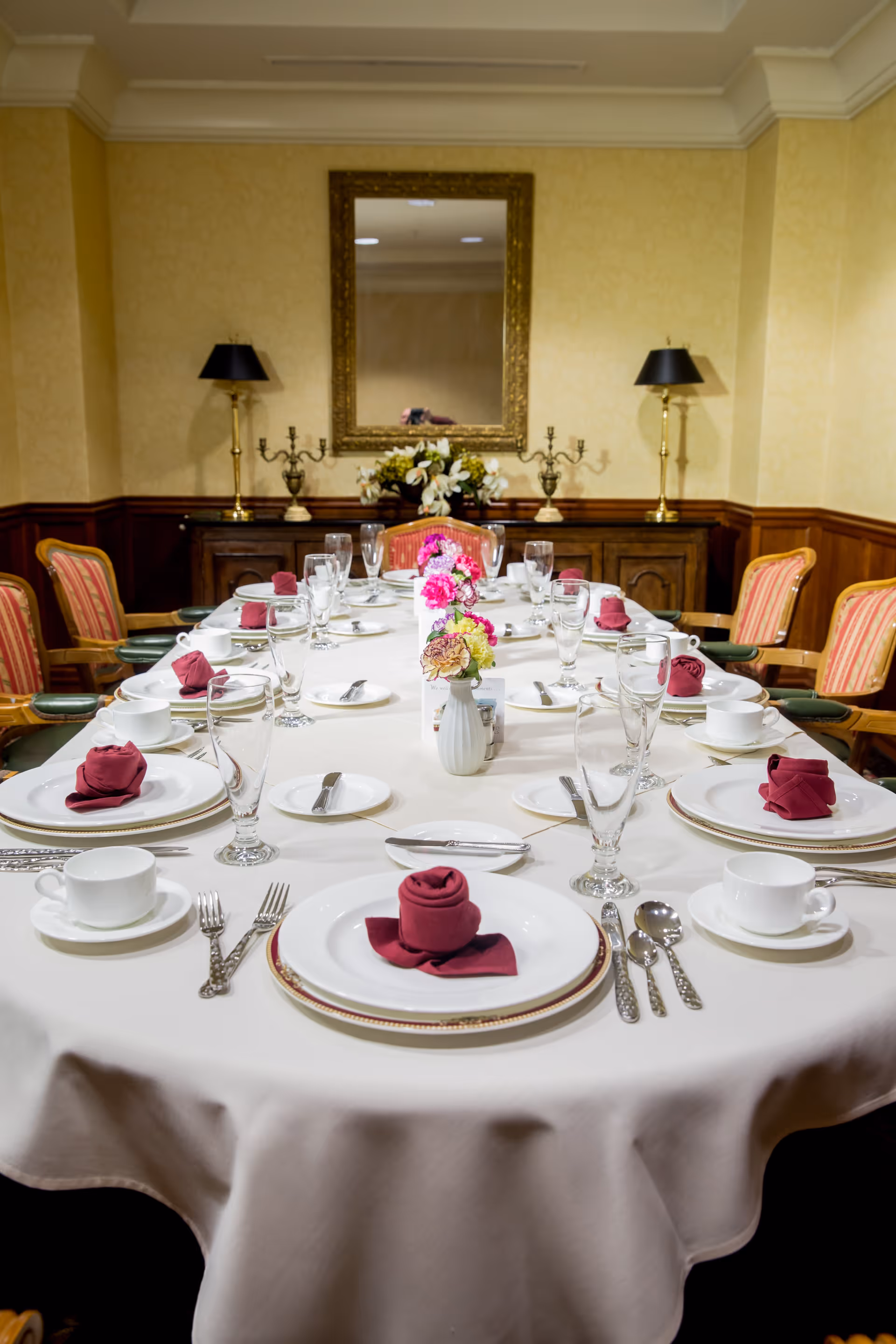 A formal dining room table set for a meal with white tablecloth, white plates, cups, silverware, and burgundy folded napkins. The table has glassware and small flower arrangements in vases. The room has yellow walls, a large mirror, two table lamps, and wooden chairs with striped upholstery.