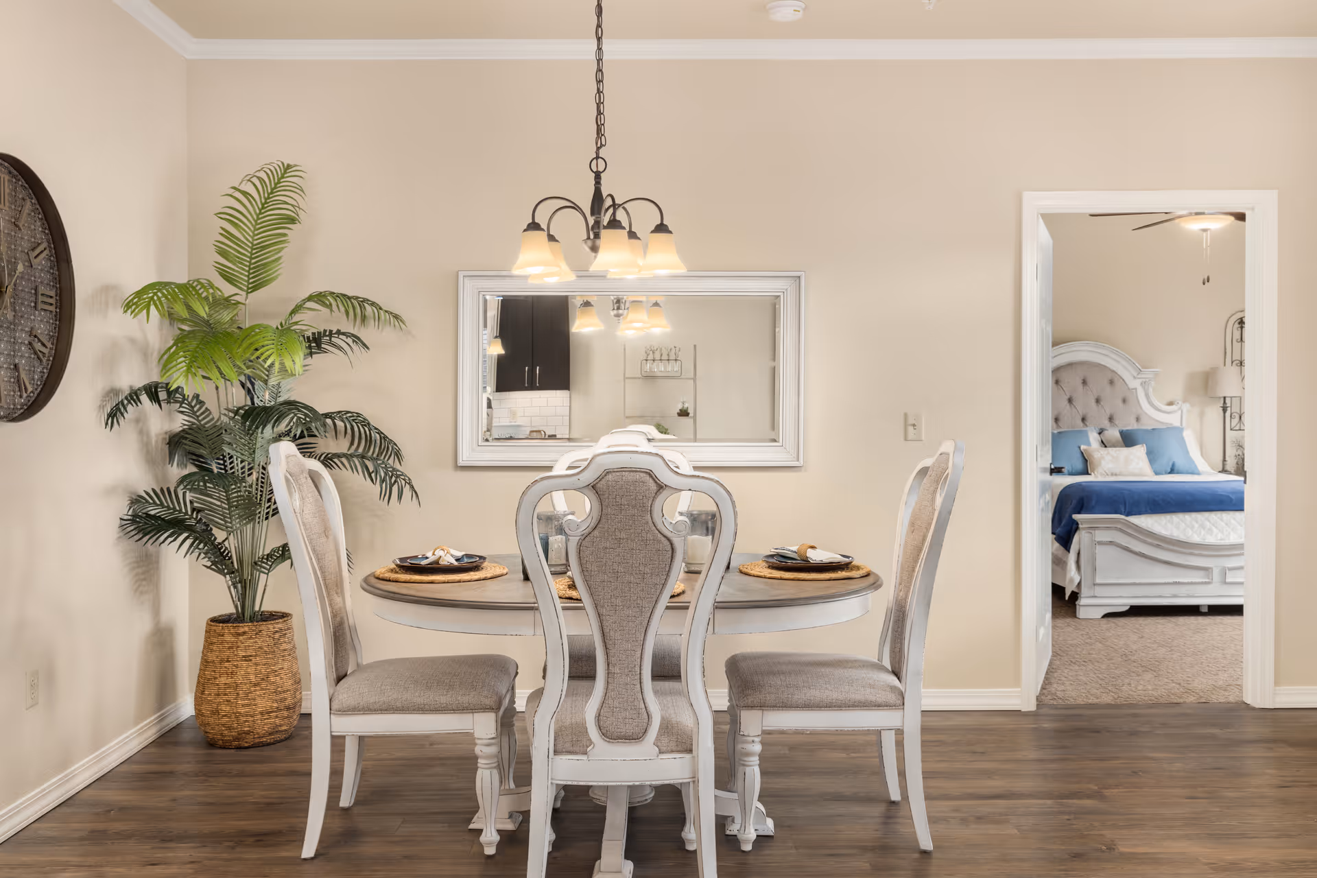 Bright dining area with a round table and four upholstered chairs under a chandelier, a potted plant to the left and a view into a bedroom through an open doorway.