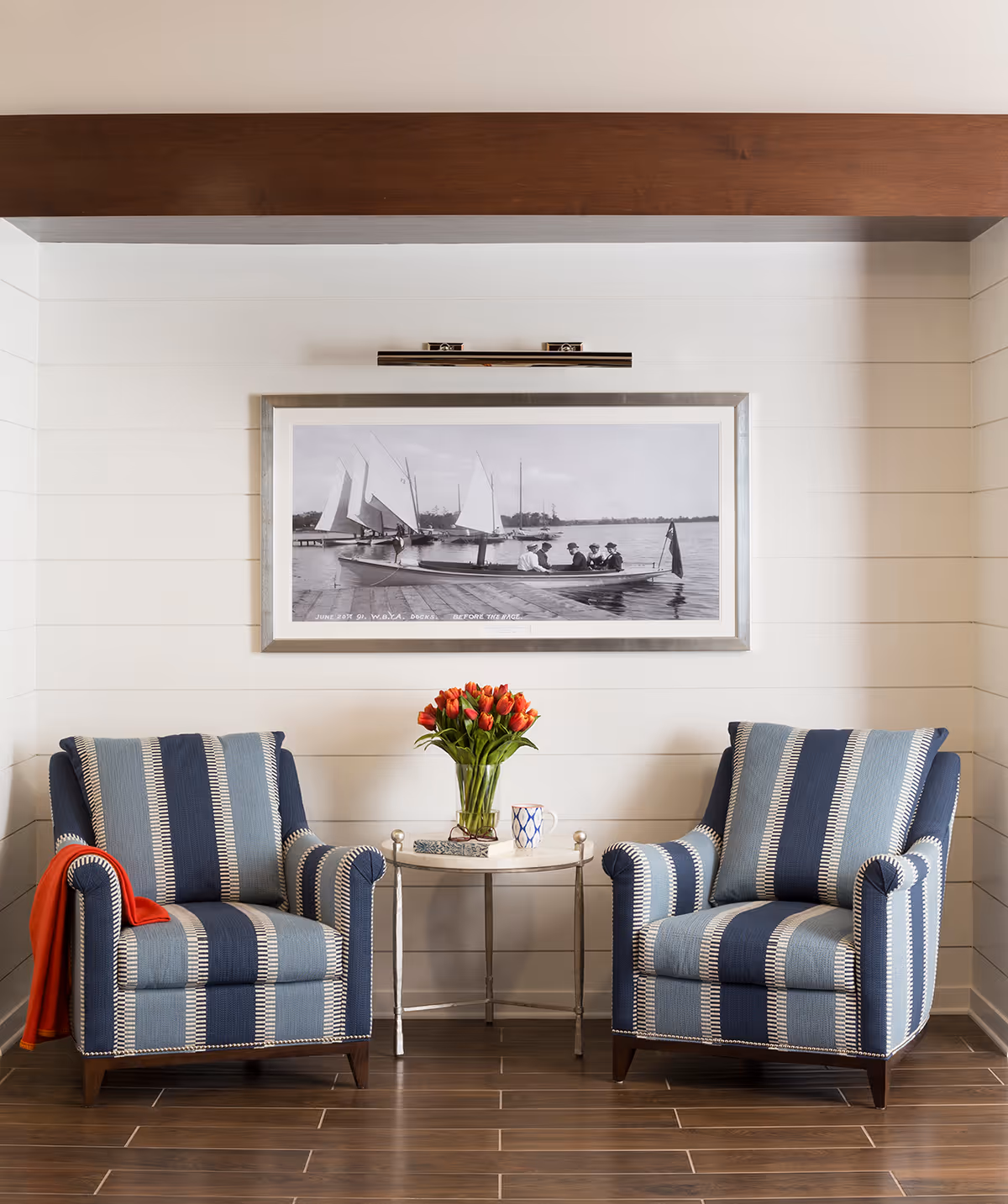 Two blue and white striped armchairs with a small round table between them holding a vase of red tulips and a coffee mug, set against a white shiplap wall with a framed black and white photo of people on sailboats above.