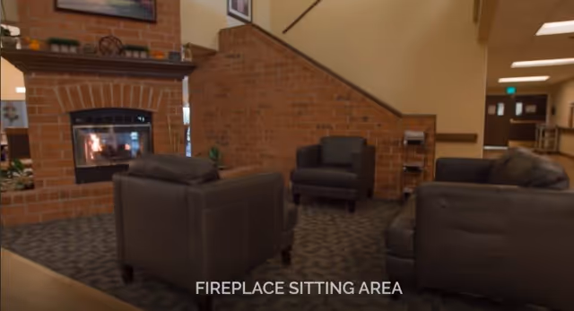 A cozy sitting area with a lit fireplace surrounded by brick walls. The space features dark leather armchairs and a sofa arranged on a patterned carpet. There are framed pictures on the walls and a staircase in the background.