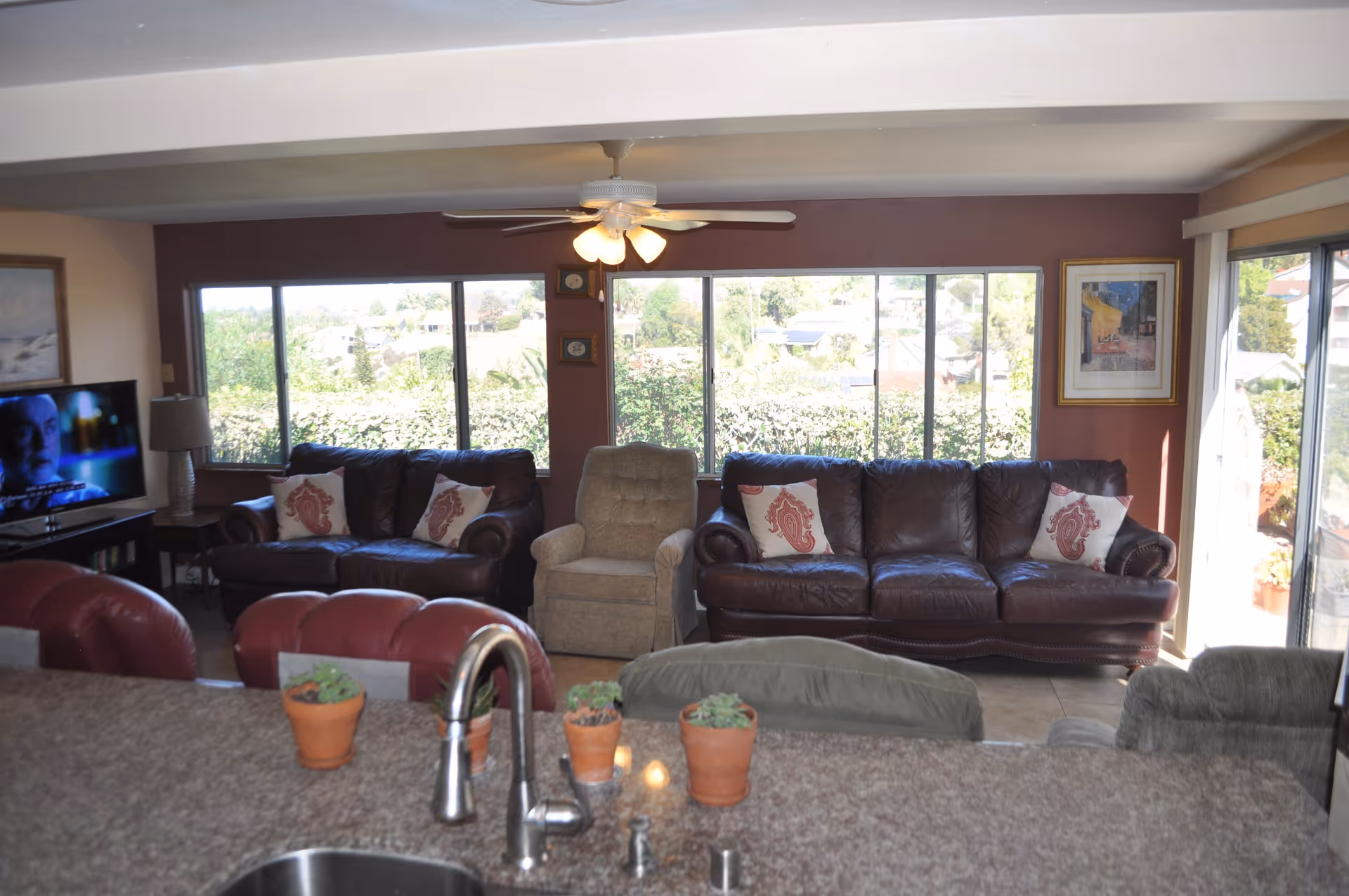 A living room area with two dark brown leather sofas, each adorned with decorative pillows, and a beige armchair in the center. Large windows behind the seating area provide a view of greenery outside. A ceiling fan with lights hangs above. In the foreground, a kitchen counter with a sink and small potted plants is visible. A television is on the left side of the room, and sliding glass doors are on the right, letting in natural light.