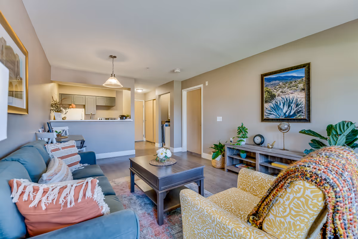 Bright furnished living room with a sofa, patterned armchair, coffee table, TV console and an open view into the kitchen.